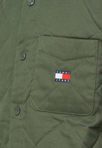 Prešívaná olivovo zelená košeľa s jacketom a čiernymi gombíkmi, s ľavým vreckom na hrudi a modrou, bielou a červenou značkou Tommy Jeans.