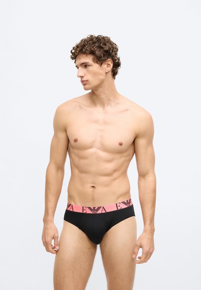 Cuecas pretas com uma cintura elástica rosa com um logótipo. O tecido parece suave e o design é justo ao corpo. Modelo está de lado.