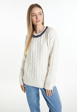 Donna con capelli lunghi e lisci che indossa un maglione a trecce bianco con righe blu navy sul collo e jeans blu, che guarda di lato.