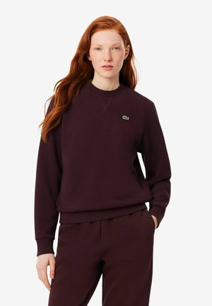 Donna con lunghi capelli rossi indossa una felpa viola scuro e pantaloni abbinati, in piedi con una mano in tasca su sfondo bianco.