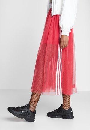Jupe en tulle rouge avec des rayures blanches, associée à des bas résille noirs et des chaussures de sport noires, mettant en valeur des textures et des couleurs superposées.