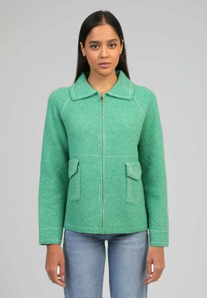 Femme portant une veste en laine verte à fermeture éclair avec poches avant et un jean bleu clair, debout devant un fond gris uni.
