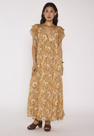 Femme debout, portant une robe longue jaune à fleurs avec des manches courtes à volants et des sandales compensées marron, sur un fond uni clair.