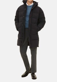 Svart pufferjacka med huva, med en quiltad design, stora knappar och en avslappnad passform, bärs över en blå tröja och grå byxor.