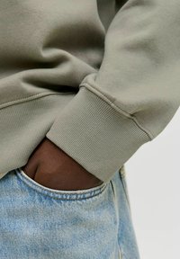 Olivzaļa sporta krekla audums ar ribotu aproci; kopā ar gaiši ziliem džinsiem, rokai kabatā. Mīksta tekstūra un ikdienišķs dizains.