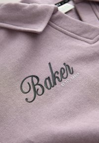 Haină mov deschis cu inscripția „Baker by Ted Baker” imprimată pe material sub un guler pliat.