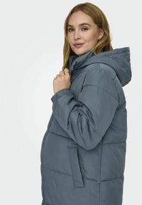 Doudoune grise matelassée avec capuche, comportant des sections matelassées, des poignets élastiques et des poches latérales. Le tissu a l'air lisse et doux.