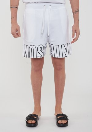 Personne portant un short blanc avec un texte noir "MOSCHINO", une chemise blanche, des sandales noires slides et des tatouages visibles sur les bras, debout.