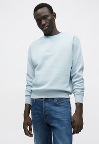 Gaiši zils džemperis ar apaļu kakla izgriezumu, ribotu aproču malu un nelielu baltu logotipu uz krūtīm, kopā ar zilām džinsa biksēm.