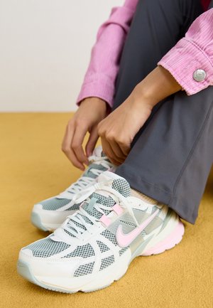 Osoba w szarych spodniach i różowej kurtce wiąże sznurówki w białych i szarych butach Nike z różowymi akcentami na żółtej podłodze.