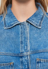 Light blue denim jacket featuring a classic collar, visible stitching, and chest pockets. The fabric has a slightly textured finish.