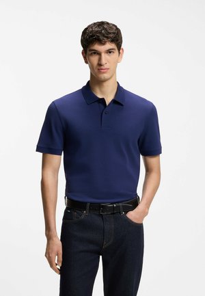 Young man wearing a navy blue short-sleeve polo shirt and dark jeans, standing with one hand in pocket against a white background.