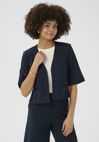 Blazer corto a maniche corte di colore navy realizzato in tessuto liscio, con scollo aperto, senza chiusura e due tasche inserite. Design semplice.