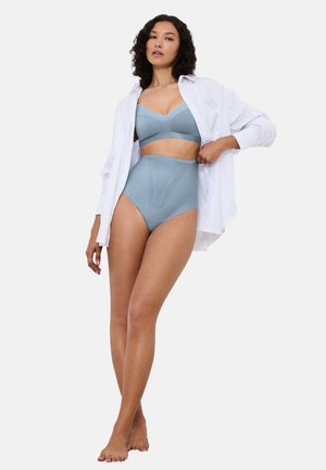 Žena stojící bosá, oblečená v světle modré podprsence a kalhotkách s vysokým pasem, bílá otevřená košile, na jednoduchém světlém pozadí.