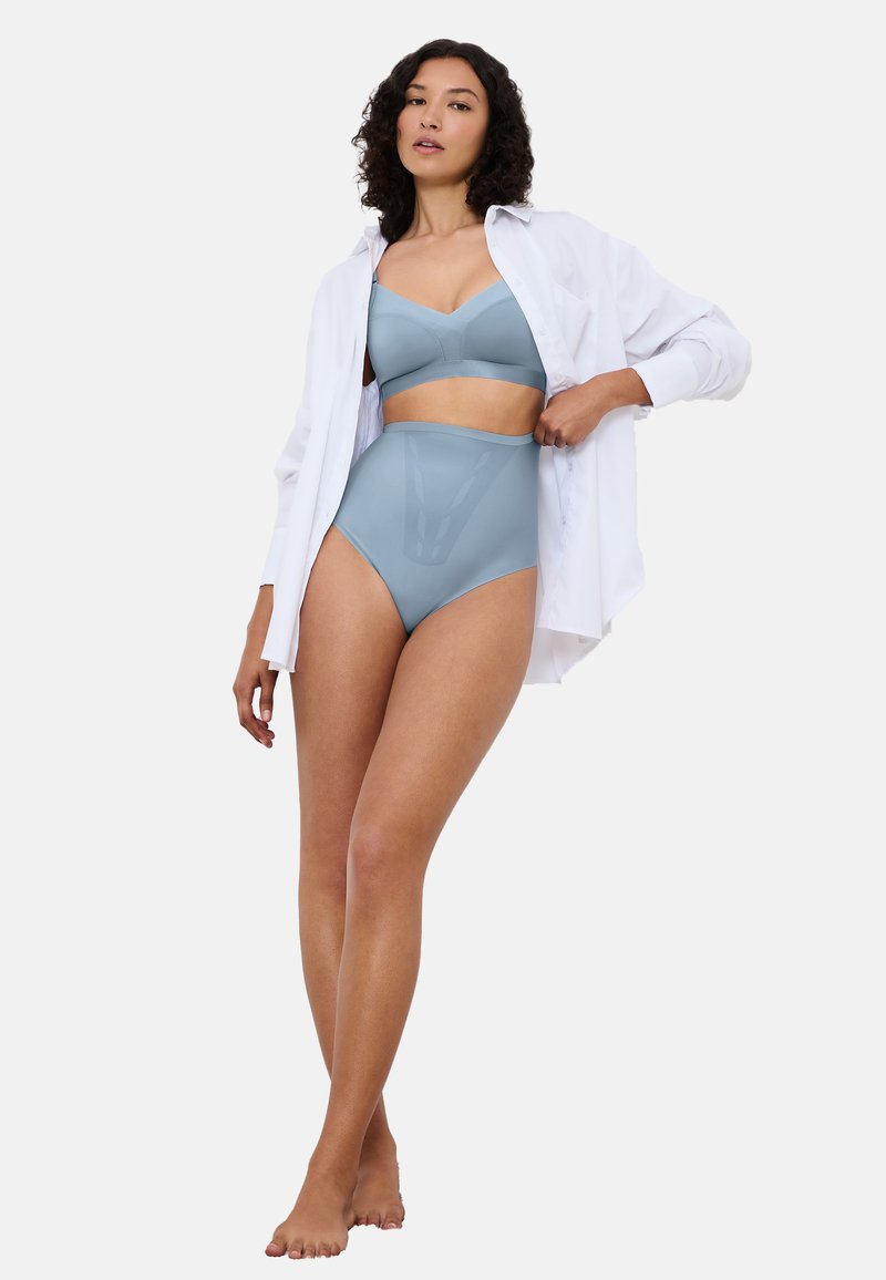 Kvinna som står barfota i ljusblå behå och höga underkläder, vit öppen skjorta, mot en enfärgad ljus bakgrund.