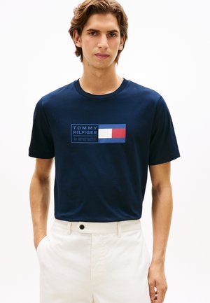 Mężczyzna ubrany w granatowy t-shirt Tommy Hilfiger z logo i białe spodnie na jednolitym białym tle.