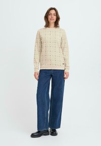 Sudadera beige con un patrón de diseños en forma de estrella negra, cuello redondo, puños y dobladillo acanalados. Combinada con jeans de mezclilla azul de pierna ancha y botas negras.