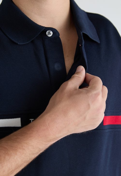 Polo shirt blu navy con colletto a bottoni, caratterizzato da un tessuto testurizzato. Strisce orizzontali rosse e bianche vicino al fondo.