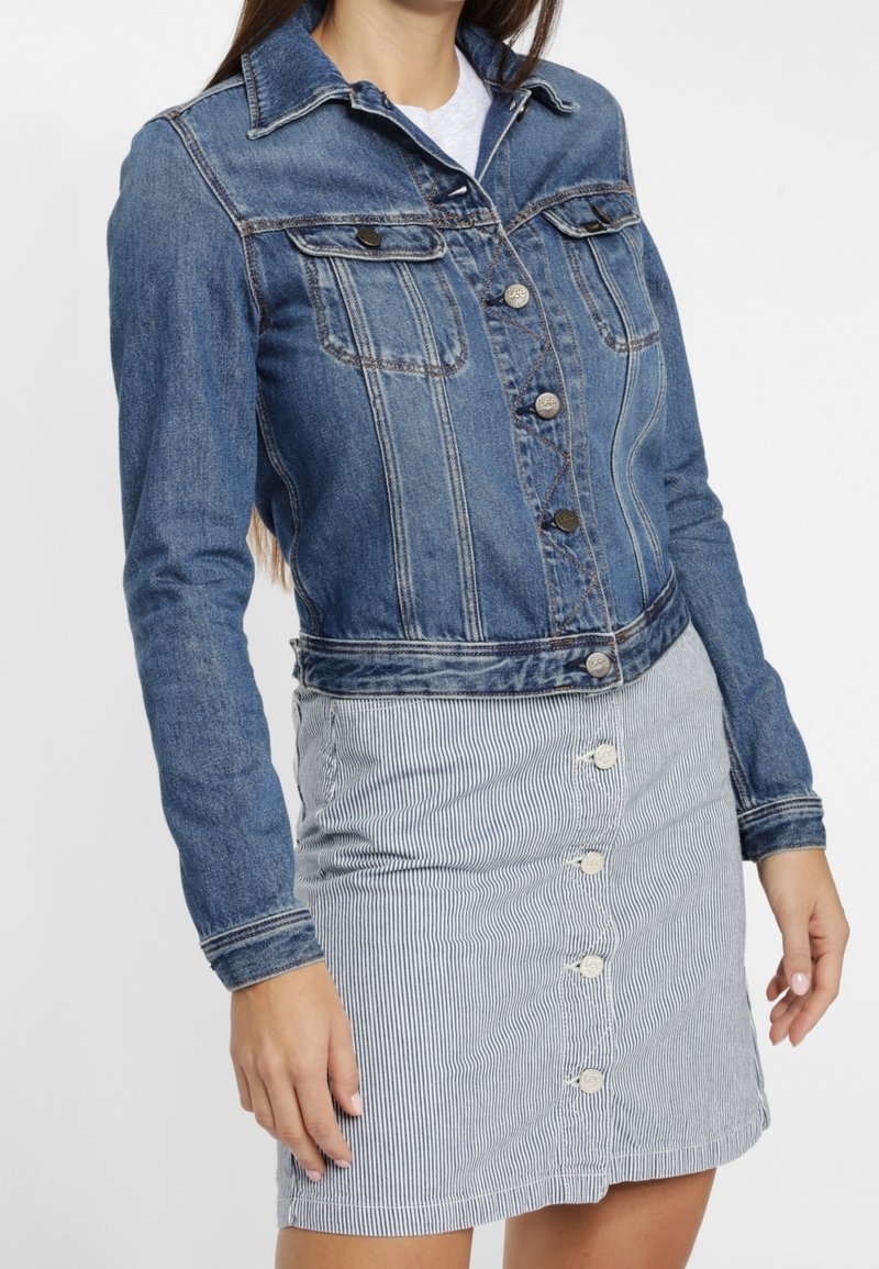 Veste en denim à la coupe ajustée, avec fermeture boutonnée à l'avant, deux poches poitrine et manches longues. Portée avec une jupe boutonnée à rayures verticales.