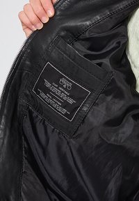 Svart läderjackas insida som visar en slät yta, silverdragkedja och en skötseltyp som innehåller produktinformation i vit text på en svart bakgrund.