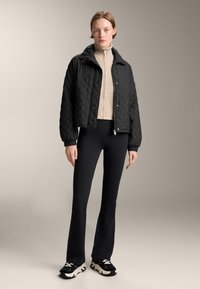 Giacca nera trapuntata con design corto, chiusura frontale con bottoni a pressione e colletto alto, abbinata a un dolcevita beige e pantaloni neri a zampa.