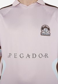 Šviesiai rožinė polo marškinėliai su trumpomis rankovėmis, turintys rudą apykaklę, išsiuvinėtą logotipą ir dekoratyvinį raštą per krūtinę.