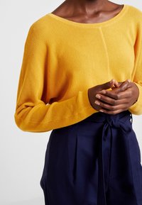 Yellow knitted sweater with a wide neckline and loose sleeves, paired with dark blue trousers featuring a tied waist.