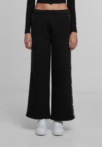 Črne široke trenirke s vrvico v pasu, ki imajo kontrastne bele stranske črte in mehko, teksturirano tkanino. Nosi se z belimi športnimi copati.