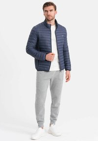 Giacca puffer blu marina con motivo a righe orizzontali, zip completa, colletto alto, abbinata a pantaloni tuta grigi chiari e sneakers bianche.