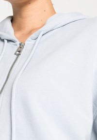 Sudadera con cremallera de color azul claro, hecha de suave tela de algodón, con cremallera plateada y logo bordado en el pecho. Cuenta con capucha y puños de canalé.