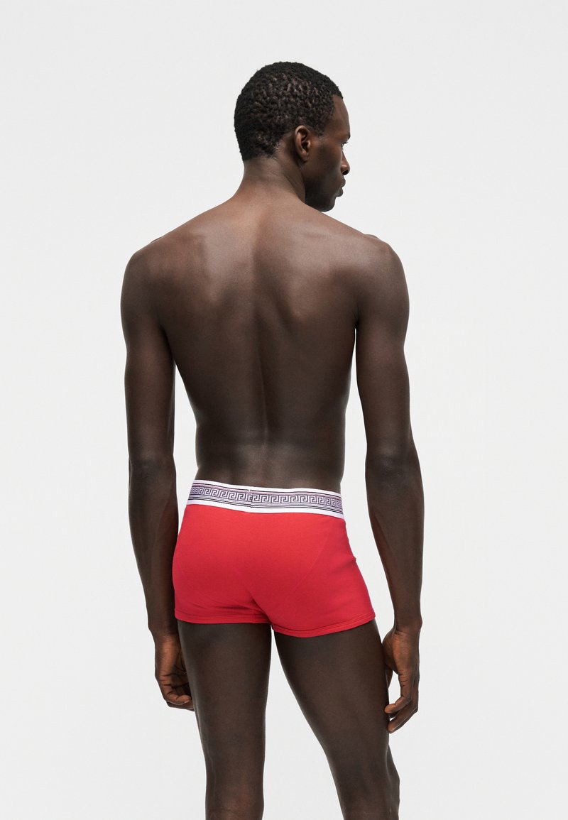 Jonge zwarte man staand met de rug naar de camera, gekleed in rode boxershorts met een witte tailleband met een geometrisch patroon.