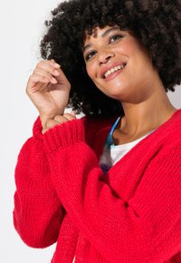 Cardigan lavorato a maglia rosso con polsini a coste, vestibilità over e superficie texturizzata. Indossato sopra una camicia chiara con un colletto blu.