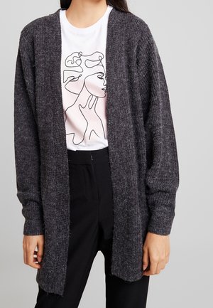 Kvinde iført en mørkegrå strikket åben cardigan over en hvid t-shirt med et sort linjetegnet ansigt-design og sorte bukser.