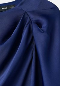 Tissu bleu marine avec une texture lisse, présentant des plis et un design plié. Étiquette du vêtement visible en haut.