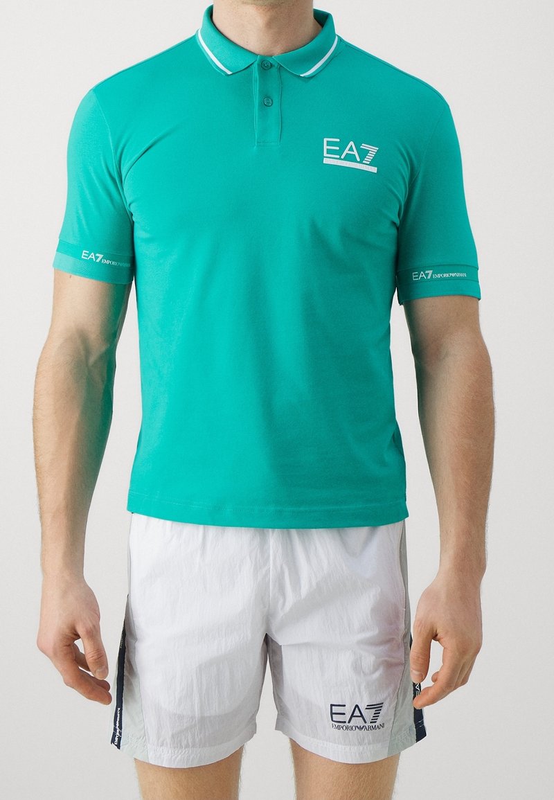 Homme portant un polo EA7 bleu sarcelle et un short de sport EA7 blanc, debout devant un fond clair uni.