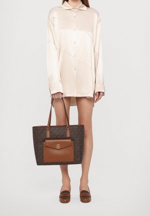 Bruine monogram draagtas met een voorvak, licht beige accenten en schouderbanden; model draagt een zijden ivoorkleurig overhemd en bruine loafers.