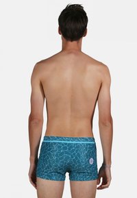 Shorts de bain pour hommes en teal avec un motif abstrait bleu clair, une taille élastique et un détail de logo sur le coin inférieur droit. Texture de tissu lisse.