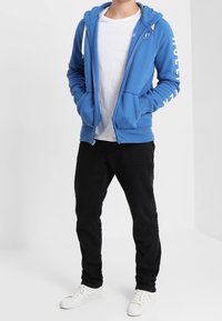 Blå zip-up hoodie med kontrasterande vit text på ärmen, ihopparad med en vit T-shirt, svarta byxor och vita sneakers.