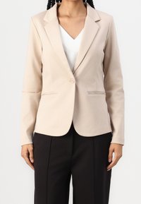 Blazer beige su misura con rever a punta, chiusura a un bottone, due tasche frontali e maniche lunghe, abbinato a una maglietta bianca e pantaloni neri.