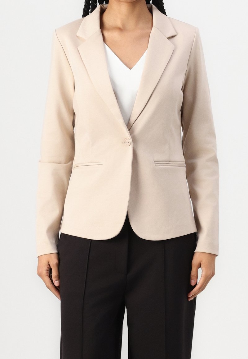 Blazer beige su misura con rever a punta, chiusura a un bottone, due tasche frontali e maniche lunghe, abbinato a una maglietta bianca e pantaloni neri.
