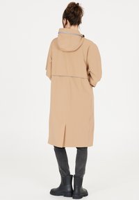 Lys beige vandafvisende jakke med hætte, korte ærmer og slids bagpå. Har grå detaljer og en afslappet pasform, parret med sorte støvler.