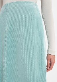 Een muntgroene rok van gestructureerd ribfluweel met diagonale ribbels, gestyled met een witte blouse met lange mouwen. Hand gedeeltelijk zichtbaar aan de zijkant.