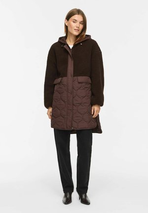 Femme portant un manteau marron à capuche avec une moitié inférieure matelassée et une moitié supérieure en teddy, pantalon noir et bottines noires, debout sur fond blanc.