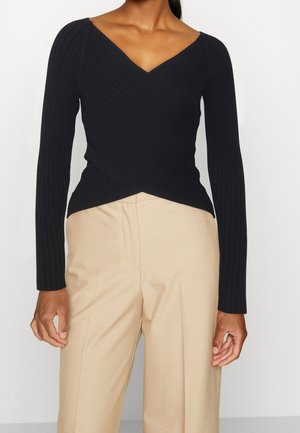 Femme portant un pull cache-cœur à manches longues côtelé noir avec col en V et un pantalon beige taille haute à jambes larges, debout devant un fond uni.