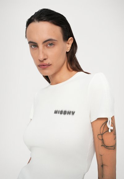 Persona con capelli scuri di lunghezza spalla, che indossa una camicia bianca aderente con il testo "MISBHV" e un tatuaggio astratto visibile sul braccio sinistro.