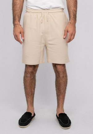 Homme portant un short beige avec cordon, une chemise blanche et des chaussures noires sans lacets, debout devant un fond gris clair uni.