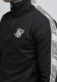 Black zip-up jacket with high collar, featuring an embroidered logo and unique white patterned side stripes. Smooth fabric and fitted design.