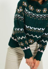 Henkilö, jolla on päällään tummanvihreä neulepaita, jossa on valkoisia, vaaleanpunaisia ja sinapinkeltaisia geometrisiä kuvioita, yhdistettynä beigeihin housuihin, kädet ristittyinä edessä.