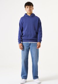 Sudadera de algodón azul con un bolsillo frontal y capucha con cordón, combinada con unos pantalones vaqueros azul claro y zapatillas blancas. Diseño simple, corte relajado.