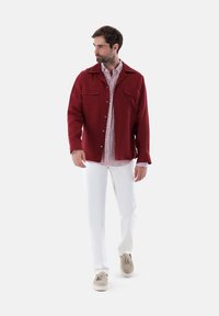 Rotes Hemdjackett mit Knöpfen und zwei Brusttaschen, getragen über einem gestreiften Hemd, kombiniert mit weißen Hosen und beigen Mokassins.